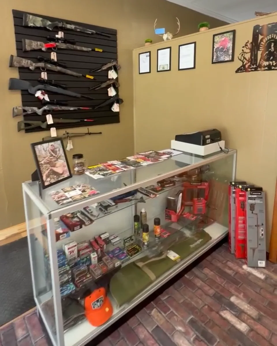 Firearms counter at Holy Ground Outdoors — long guns on the slat wall above a glass case of ammunition and reloading gear