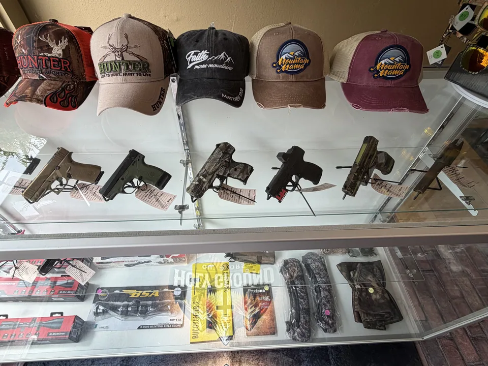 Glass display case of handguns beneath a row of hunting caps, with scopes and camo holsters